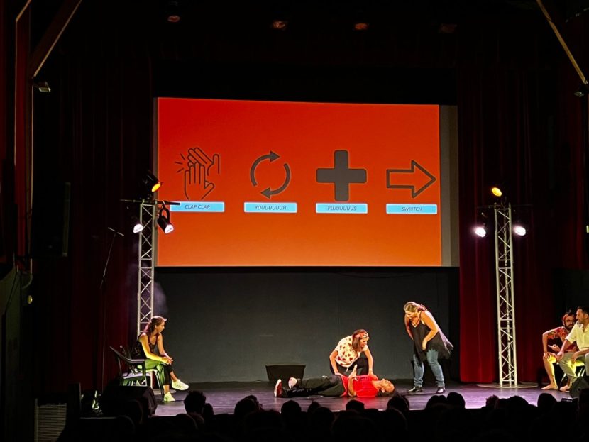 Photo d'une scène de théâtre pendant un spectacle d'improvisation. Sur le mur sont projetés quatre icônes, avec les textes "clap clap", "youuuh","pluuus" et "switch".